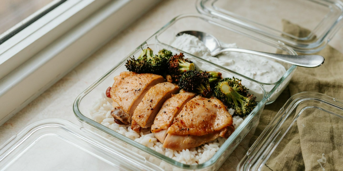 Sliced roasted chicken thighs over white rice with charred broccoli in a glass container