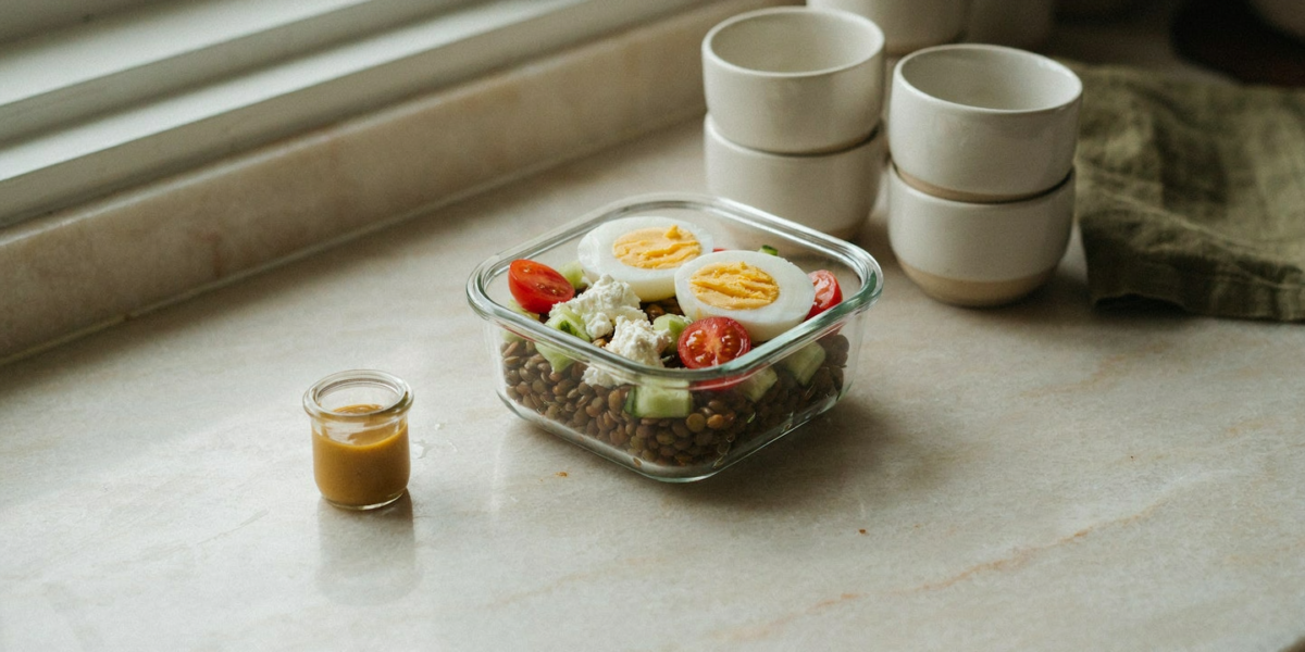 A glass bowl of lentils with halved hard-boiled eggs, feta, cucumber and tomatoes